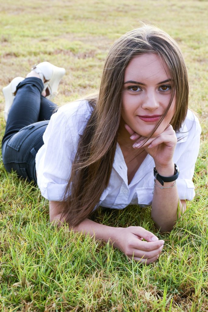 A person lies on a grassy field wearing a white shirt, dark jeans, and white shoes. They have long hair, a watch and bracelet on the left wrist, and hold a small object in the right hand. The relaxed pose and natural setting suggest a moment of rest or contemplation.