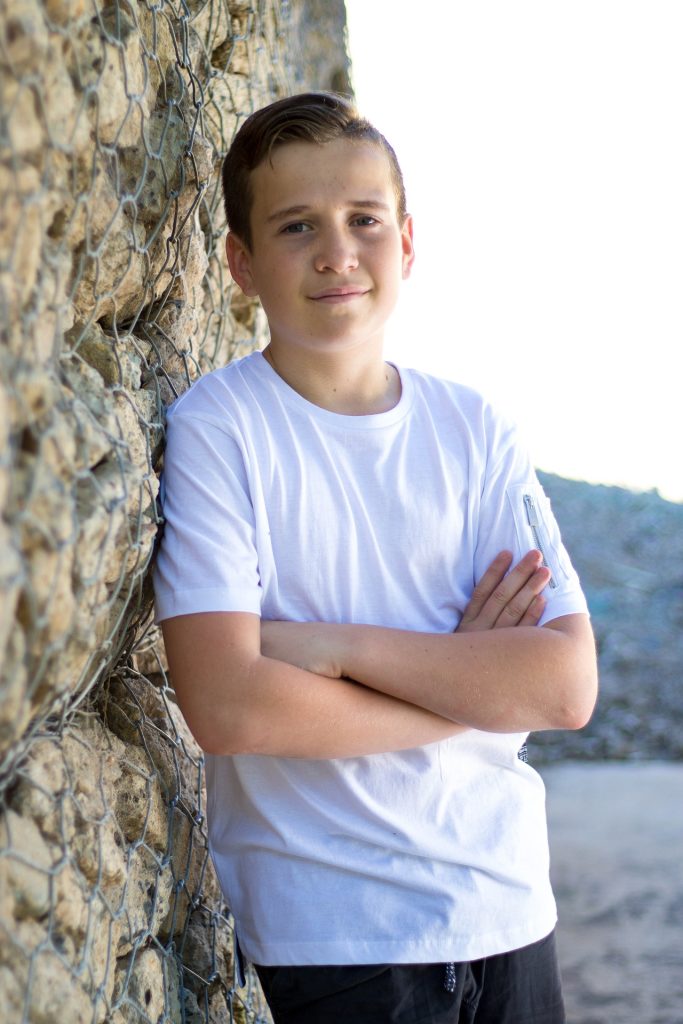 A person stands casually with arms crossed next to a stone wall reinforced with wire mesh. They wear a white t-shirt and black pants. The outdoor setting, possibly near a beach or rocky area, contrasts the smooth clothing with the rugged texture of the wall.