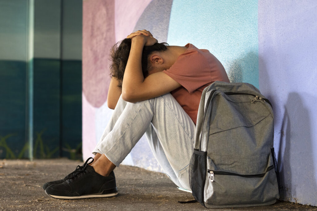A male teen sitting down next to his backpack after school; the teen is depressed and is in clear need of treatment. 