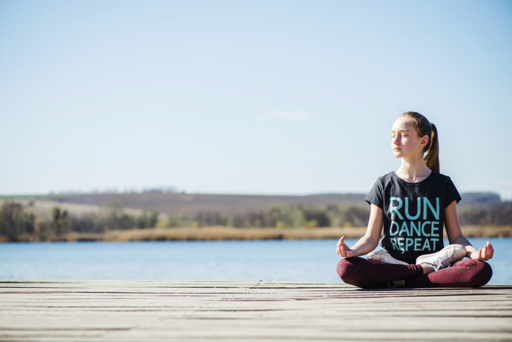 A female teen is meditating by the lake; as a mechanism to cope with any depression that might sneak up on her. 