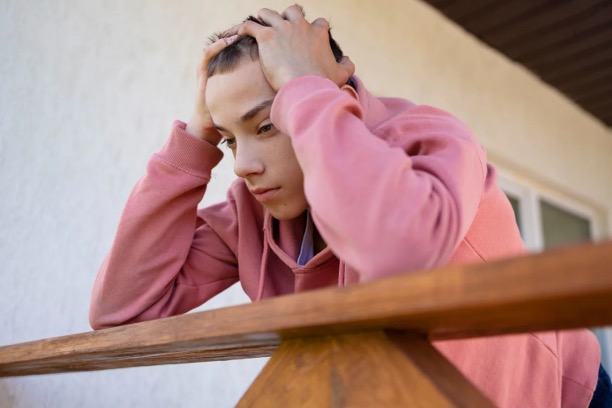 A teenage boy in a pink hoodie leans on a wooden railing outside, holding his head in his hands