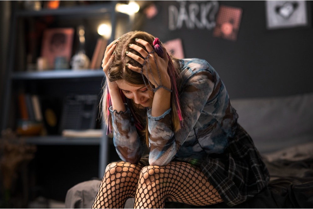 A teenage girl with pink-streaked hair sits on a bed in a dim room, head in her hands