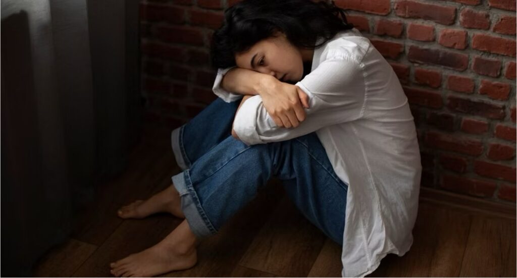 A young woman sits on a wooden floor against a brick wall, hugging her knees with her head resting on her arms.