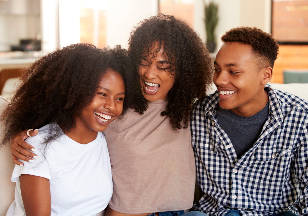 Three smiling young adults sitting close together on a couch, sharing a joyful moment—symbolizing the supportive, healing community and emotional connection fostered at a rehab center.