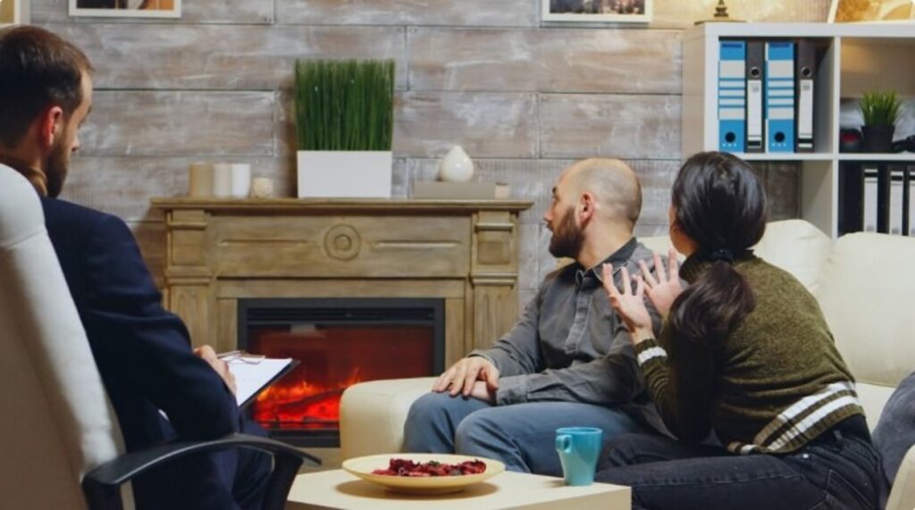 A couple sits together on a sofa during a counseling session, with the therapist taking notes on a clipboard.