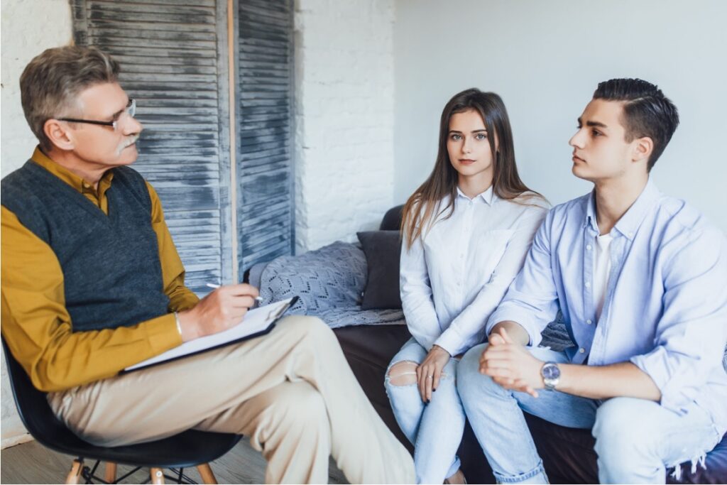 An older male therapist in glasses takes notes while a young couple sits across from him on a sofa