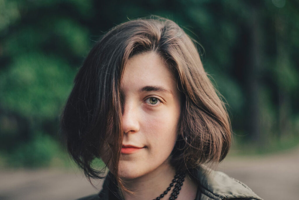 Young woman with short brown hair and a calm expression, standing outdoors with greenery in the background—symbolizing personal reflection, resilience, and the journey toward healing at a rehab center.
