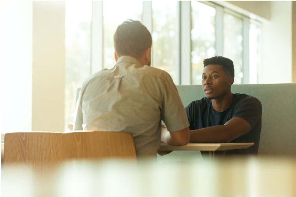 Two men engaged in a one-on-one conversation in a bright, modern setting—representing supportive counseling or therapy as part of a rehab center's personalized recovery program.