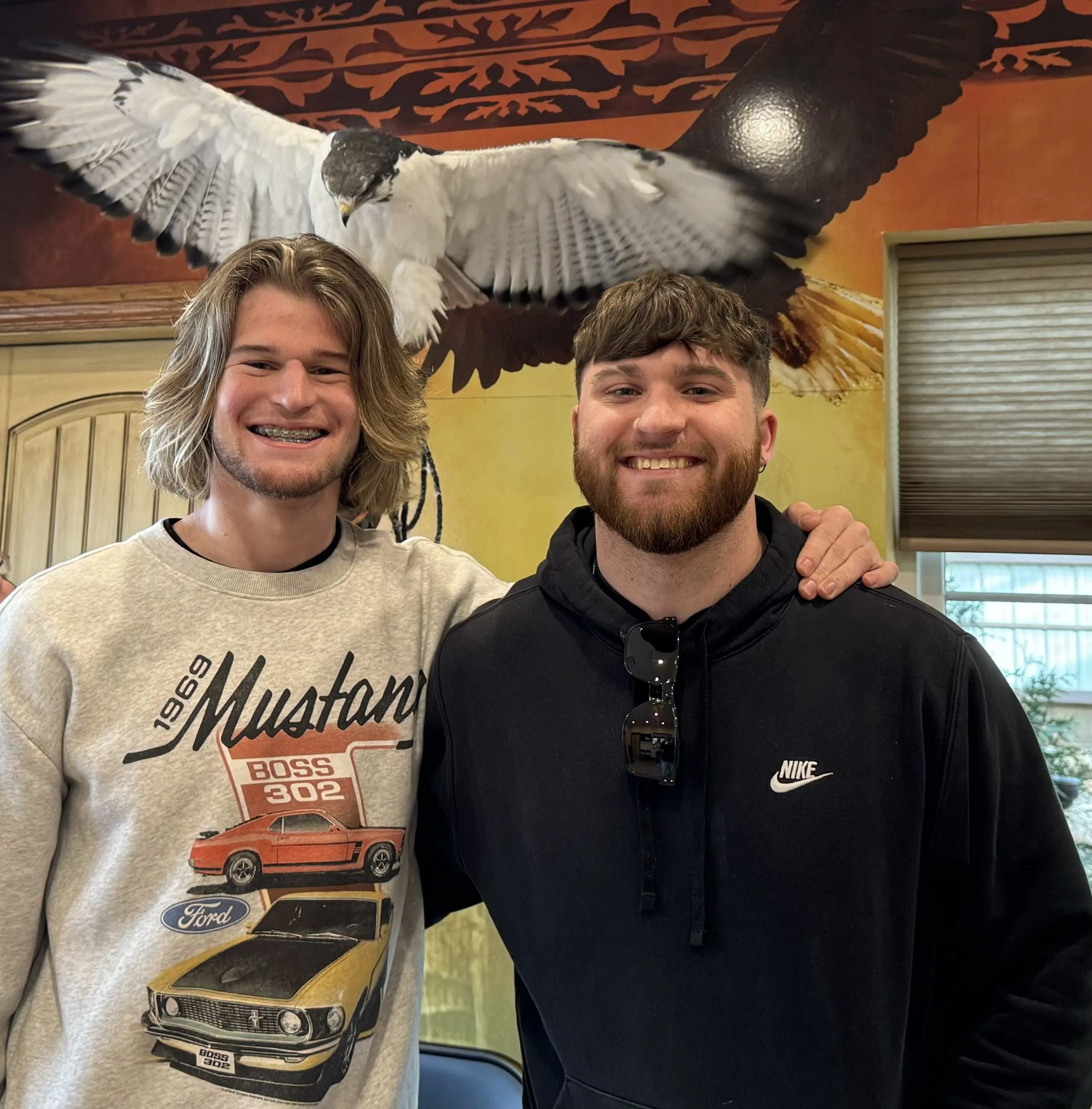 Two young men smiling and standing side by side inside a rehab center, with one resting his arm around the other’s shoulder. They are in front of a mural featuring a large bird in flight, symbolizing freedom and recovery.