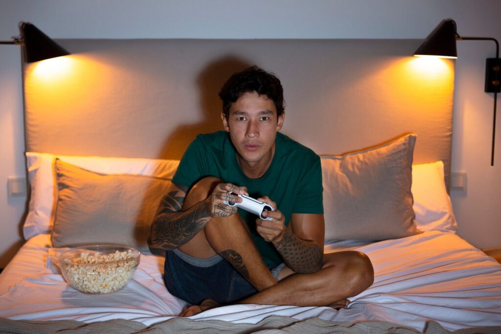 Young teen sitting cross-legged on a bed playing a video game with a bowl of popcorn beside him