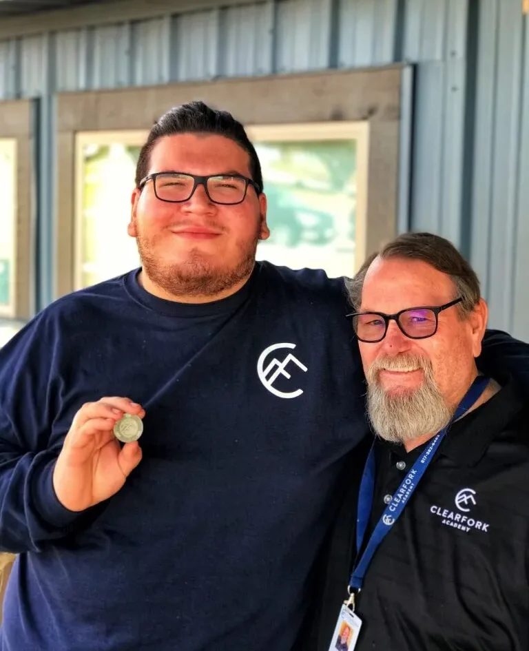 A young man celebrating a recovery milestone while proudly holding a sobriety coin, standing beside a smiling staff member from Clearfork Academy, symbolizing support and achievement in a rehab center.