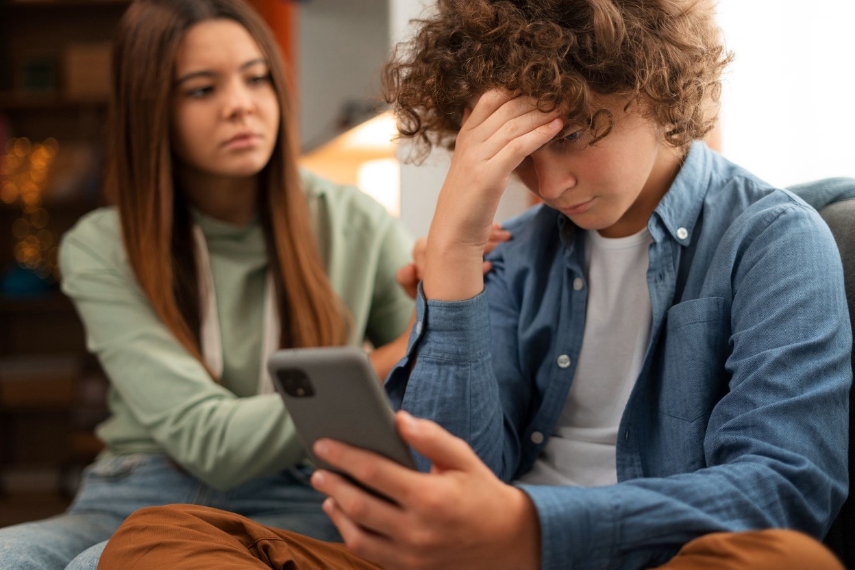 A teenage boy looking distressed at his phone while a female friend comforts him
