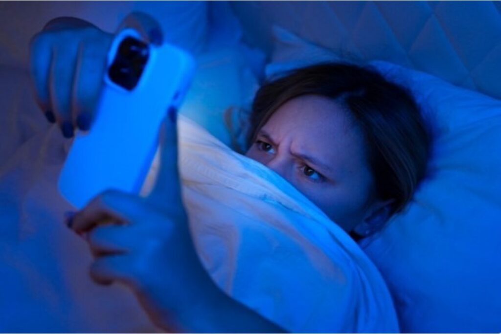 A teenager lying in bed at night, illuminated by the blue glow of her smartphone screen