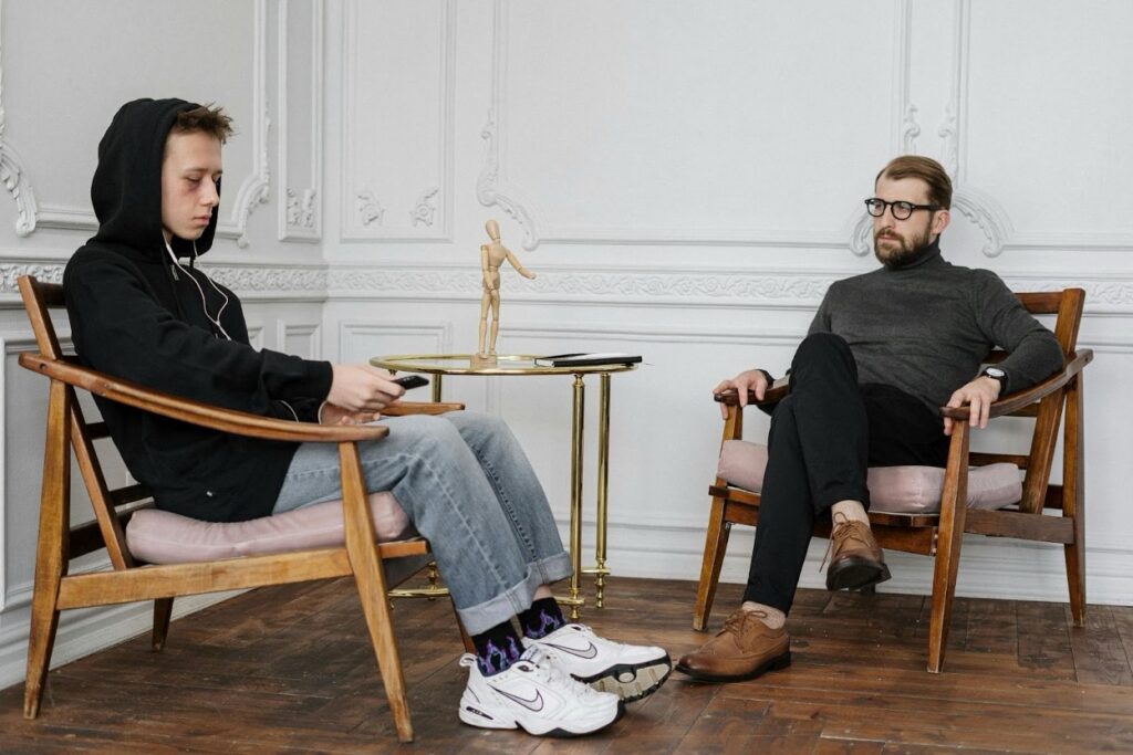 A hooded teenager looks at his phone while a bearded adult counselor watches him