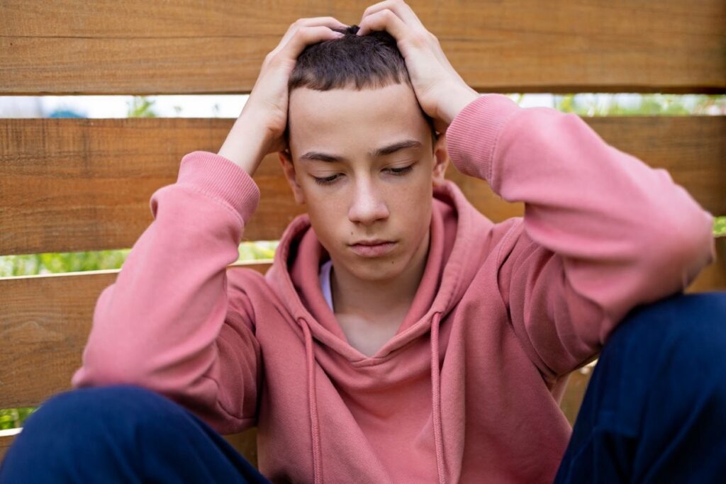 A teenage boy in a pink hoodie sits against a wooden fence in distress