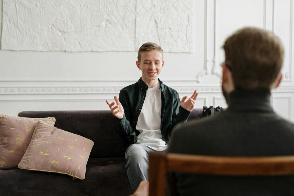 A teenage boy sits on a sofa speaking expressively to an adult man