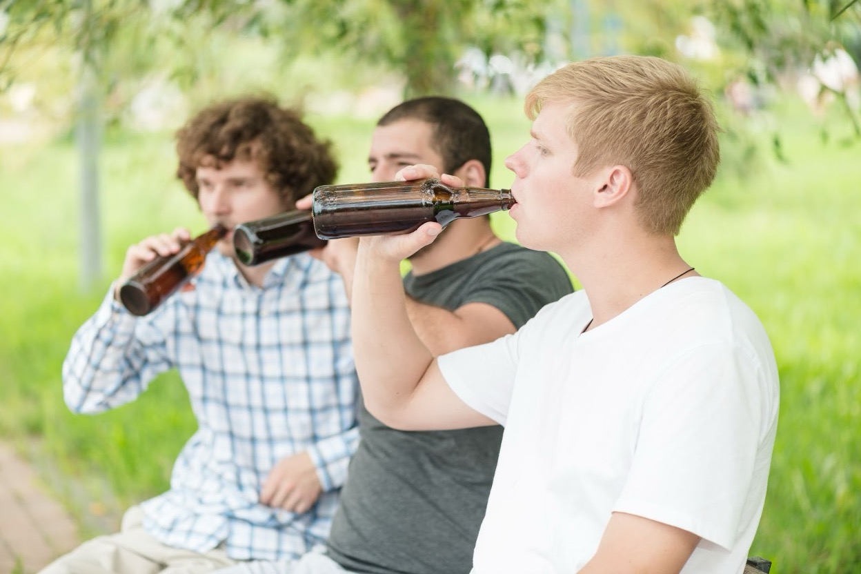 Three young men sit outdoors in a park simultaneously drinking from beer bottles