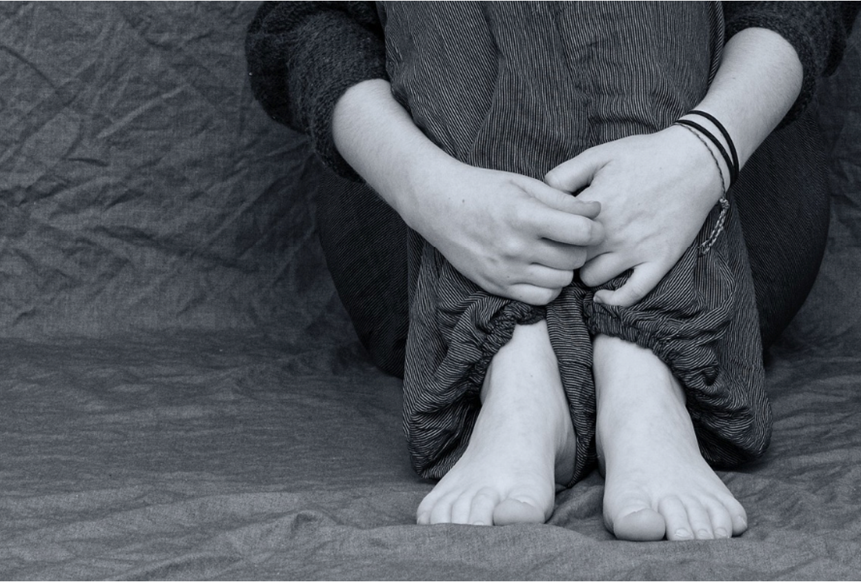 A troubled teen sitting on the floor, hands tightly holding their knees together, showing signs of emotional distress and self-harm.