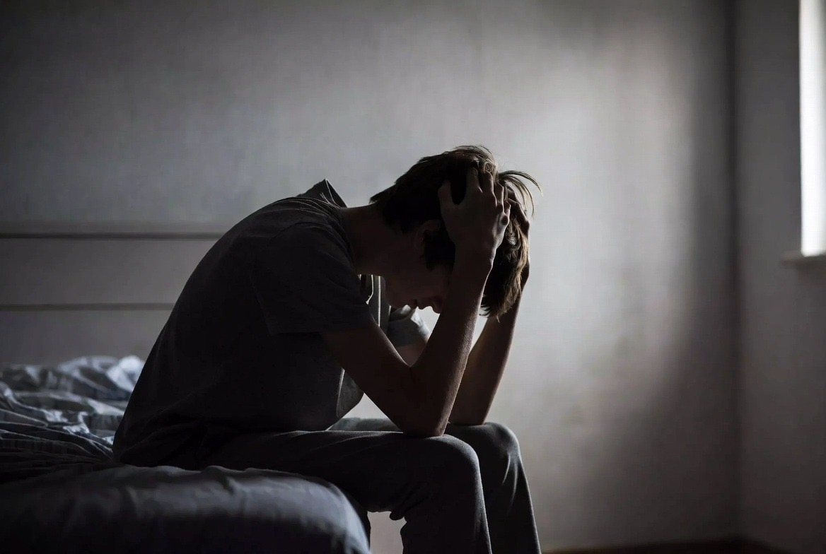 A teenage boy sitting on the edge of a bed with his head in his hands, looking exhausted and emotionally withdrawn