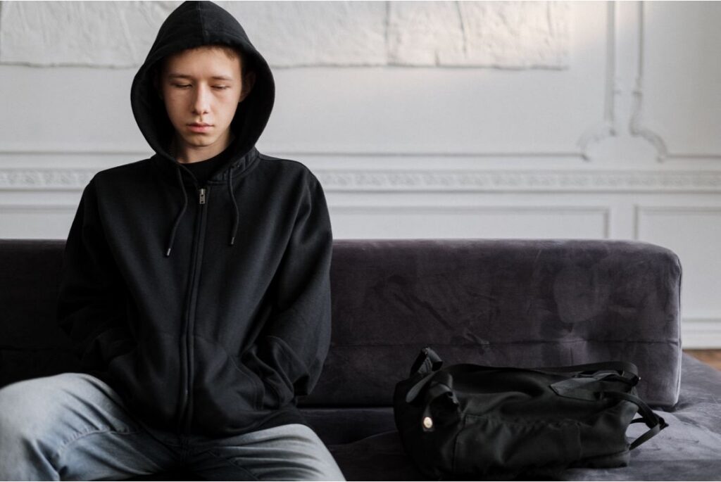 A teenager in a black hoodie sits slumped on a couch beside a dark bag.