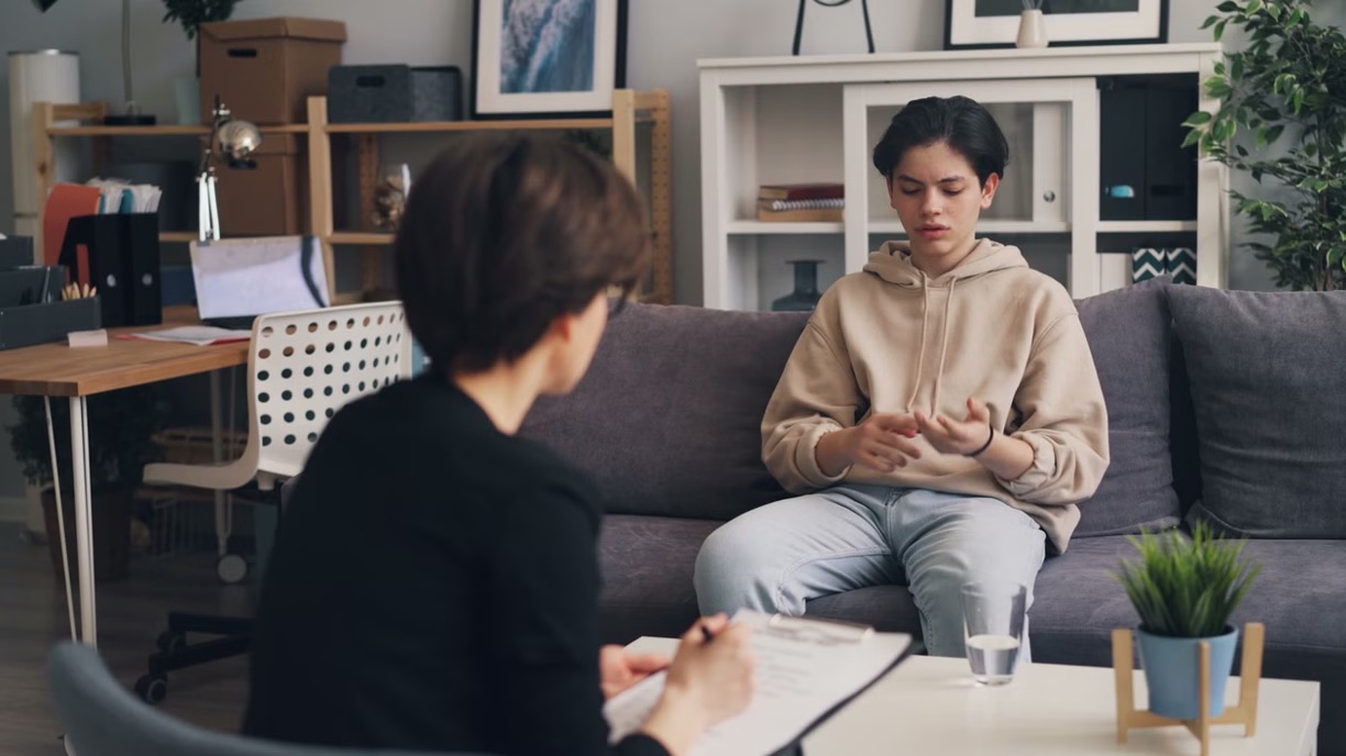 A teenager sits on a sofa talking to a therapist who takes notes on a clipboard