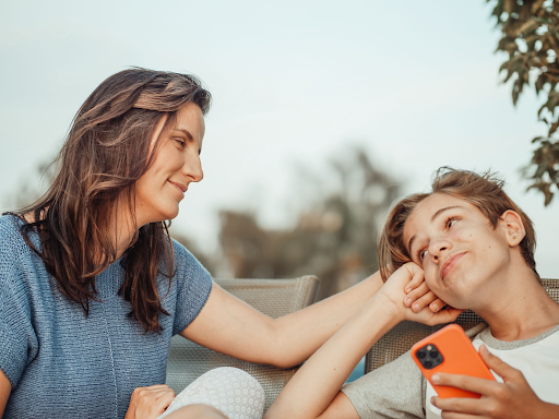 A woman and a teenage boy holding an orange phone, smiling at each other outdoors