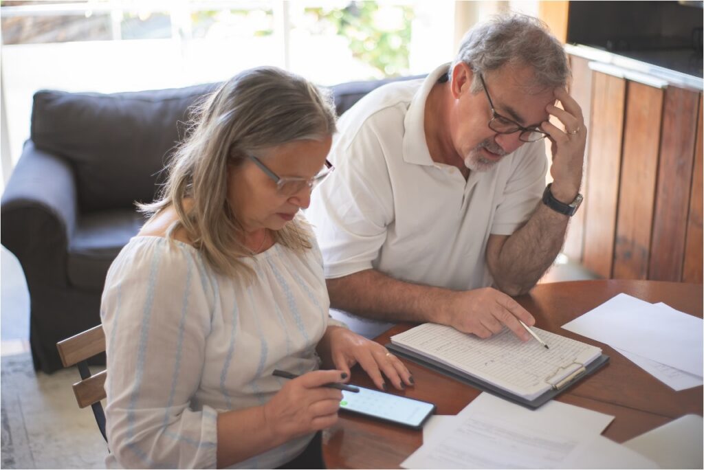 A worried older couple reviews paperwork and finances together at a table using a calculator.