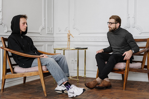 A young man in a black hoodie sits facing a bearded therapist during a counseling session