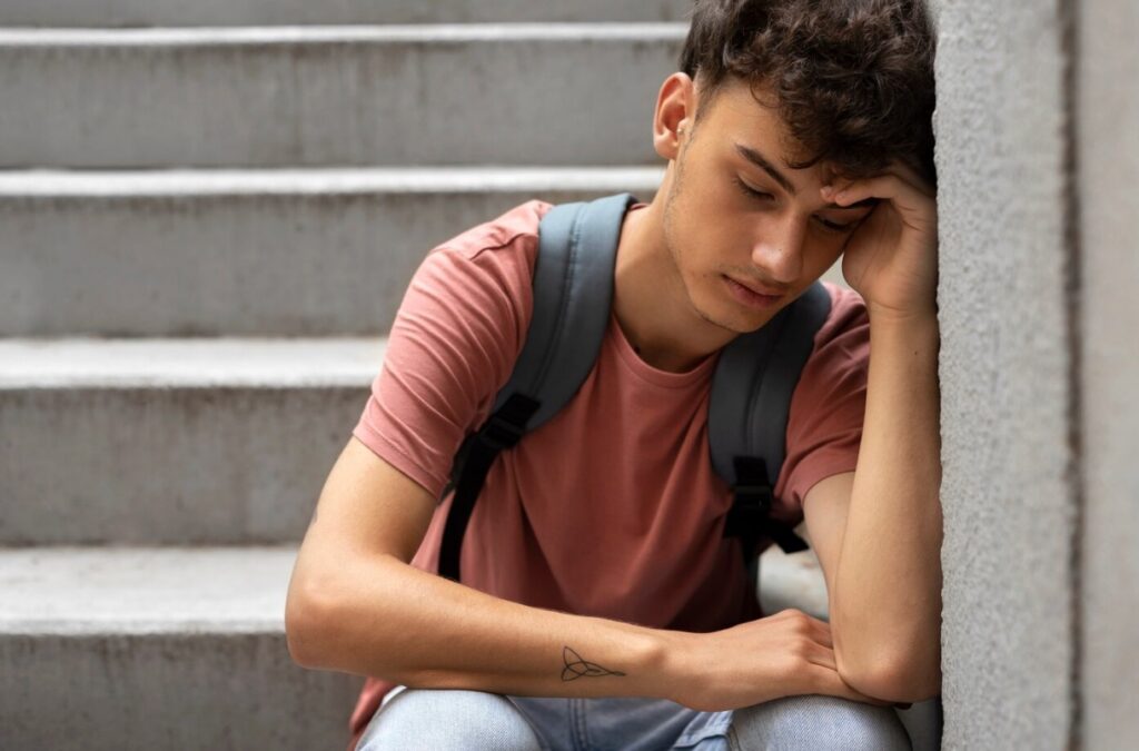 A young man with a backpack sits on concrete steps, looking down and resting against a wall.