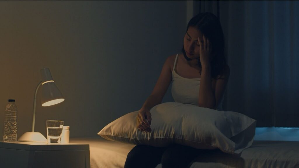 Distressed teenager sitting on the edge of a bed in a dark room, unable to sleep.