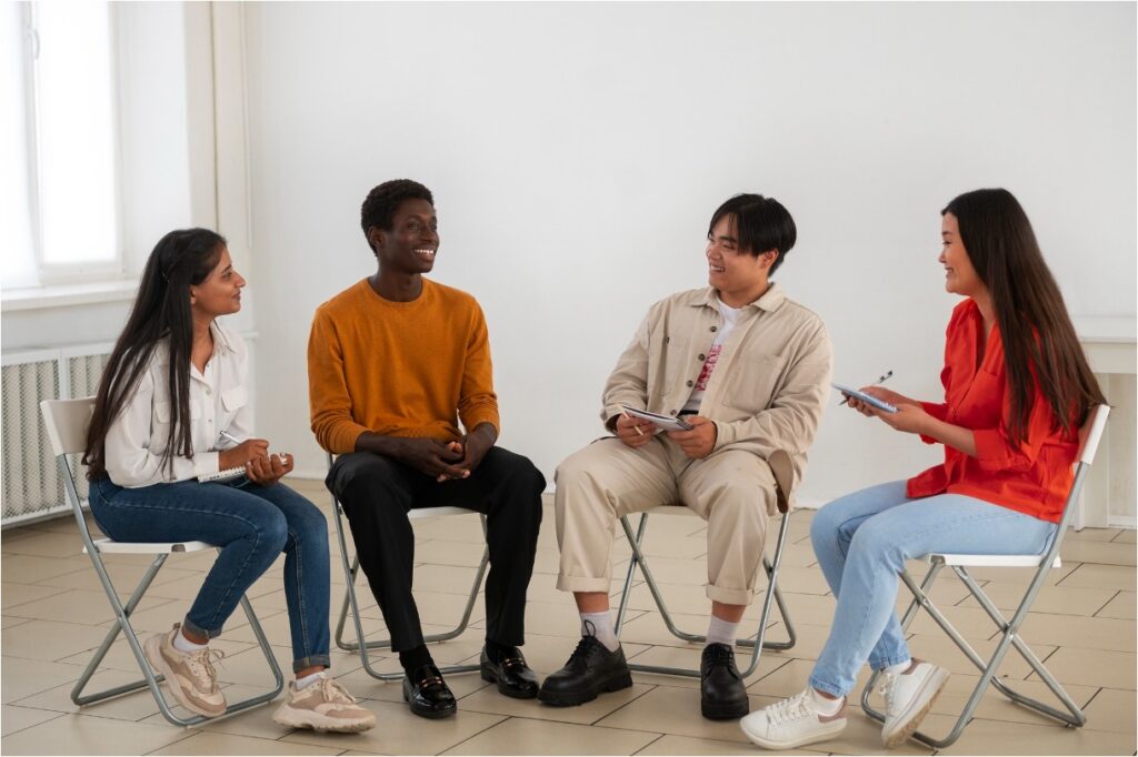 Four young people sit in a circle on folding chairs, smiling and talking during a group session.