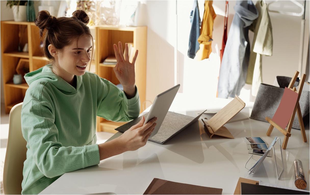 Smiling young woman in green hoodie waves at tablet during video call at desk