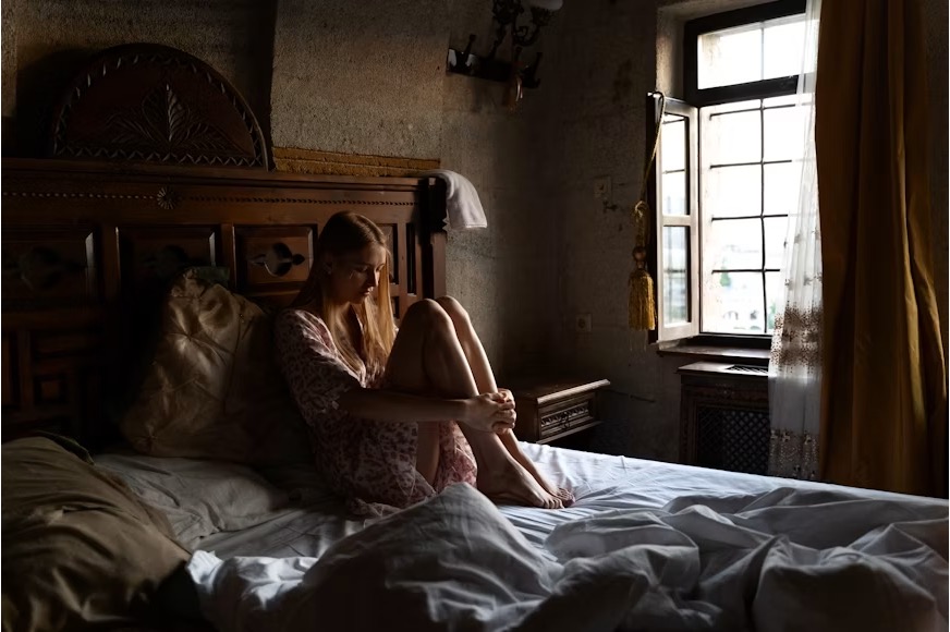 Teenage girl sitting alone in a dimly lit bedroom, looking down with a sad expression.