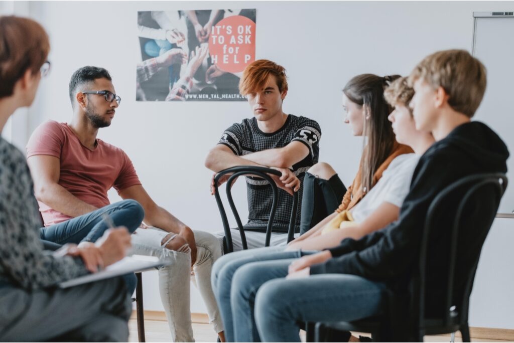 Teenagers in a group therapy session, seated in a circle, with a therapist taking notes.