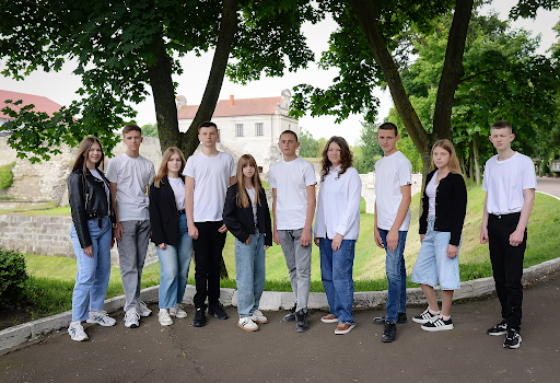 Ten teenagers in white and black outfits stand in a row outdoors near a stone building and trees