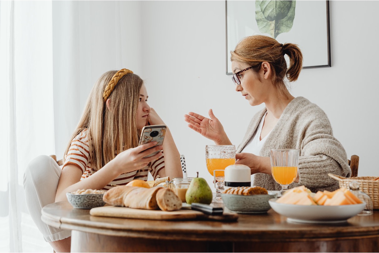 Woman gestures while talking to a disinterested teenage girl holding a phone at a breakfast table