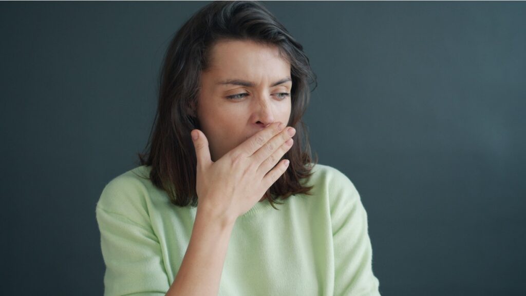 A woman in a green sweater covering her mouth, looking fatigued and nauseous against a gray background