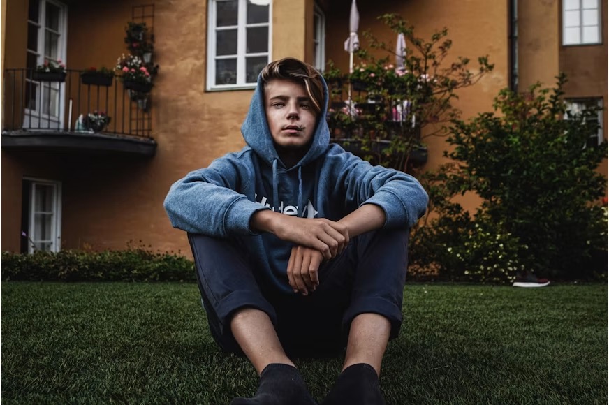 Distressed teen in a blue hoodie sitting on green grass with his arms slightly folded