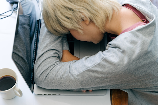 Person in a gray sweater slumped face-down on a laptop keyboard beside a cup of coffee