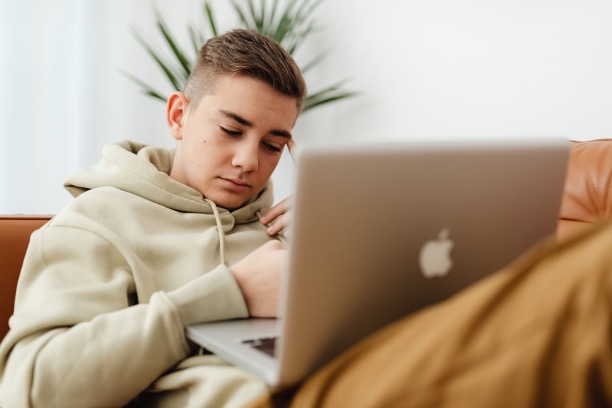 Teen in a beige hoodie sitting on a couch looking tired while using a laptop