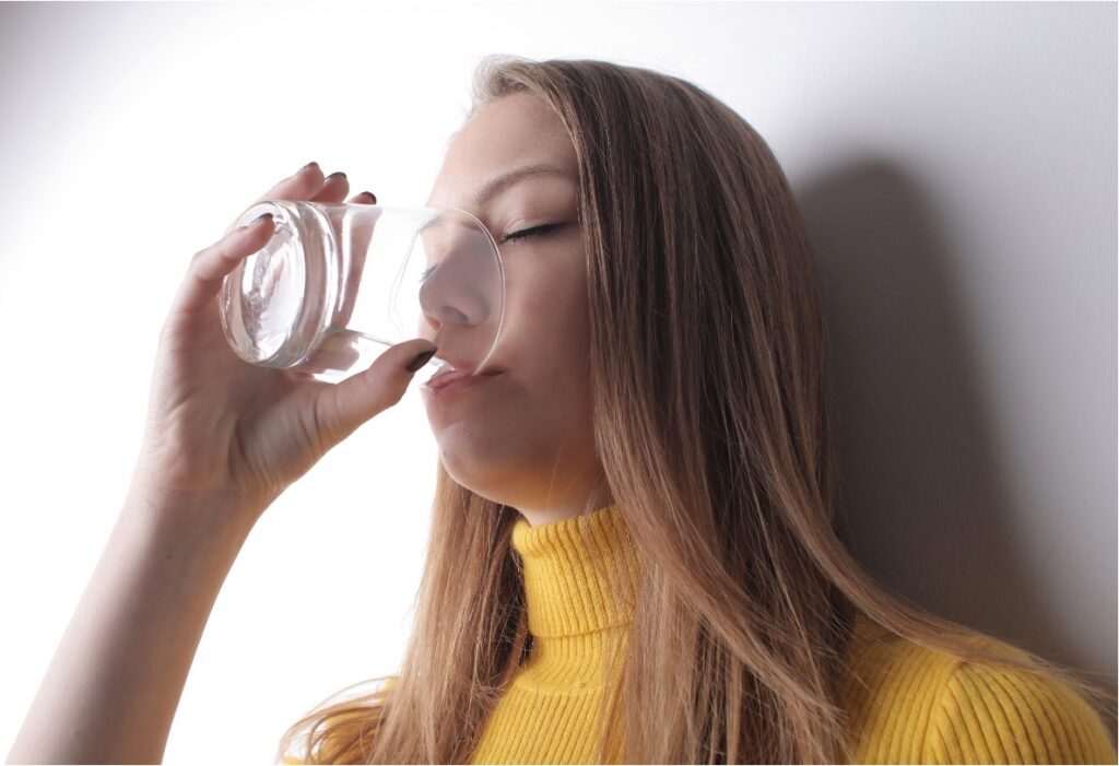 Young woman in a yellow turtleneck drinking a glass of water against a light background.