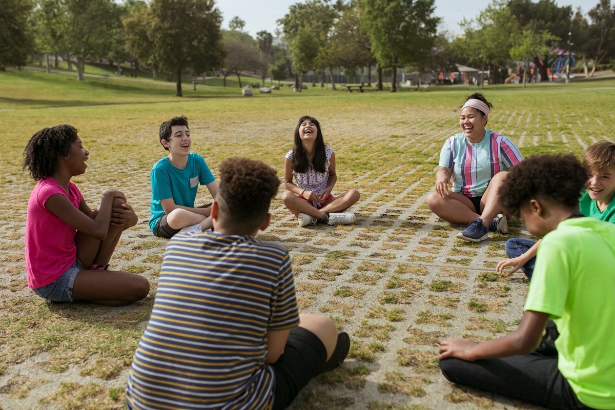 A group of teenagers laughing together while seated in a circle outdoors in a park