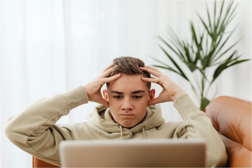 A teen in a beige hoodie holds his head while staring at a laptop, struggling to focus