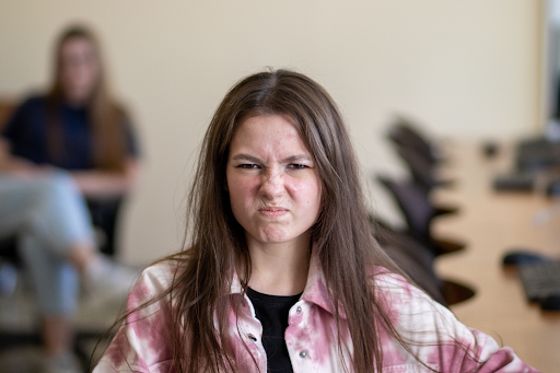 A teenage girl in a pink tie-dye shirt scowls, nose wrinkled, visibly angry, in a classroom