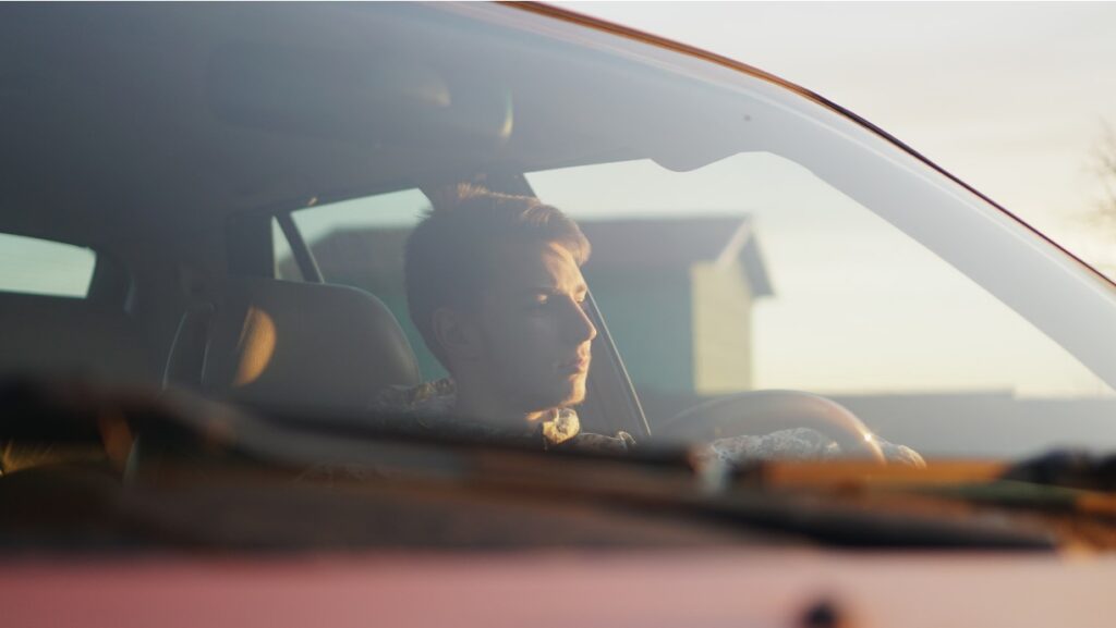 A young driver is seen through a windshield, hands on the wheel, staring ahead with a blank expression