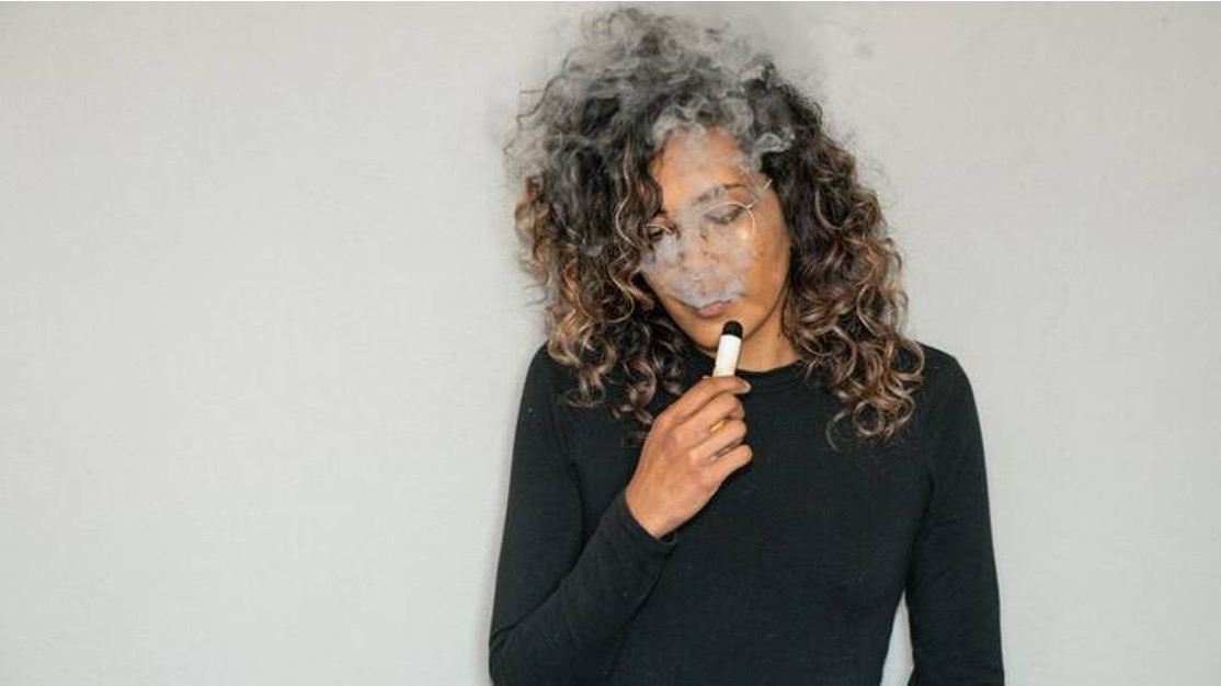 Curly-haired teen in glasses exhaling a cloud of vapor while holding a white disposable vape