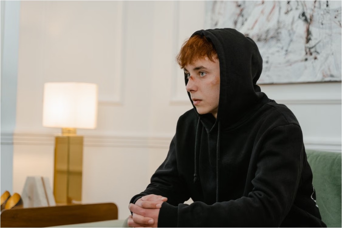 Red-haired teen in a black hoodie sits with clasped hands, looking thoughtful in a calm therapy office