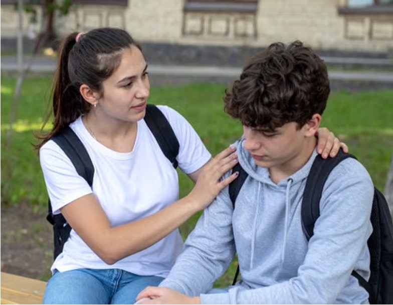 Teenage girl comforts a downcast teenage boy, hands on his shoulders, as they sit on a bench