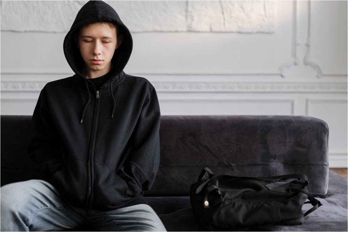 Withdrawn teenage boy in a black hoodie sits alone on a sofa, eyes downcast, backpack beside him